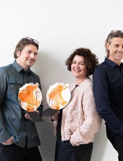 Drei Personen stehen nebeneinander, lächeln und halten kunstvolle orangefarbene und weiße Auszeichnungen vor einem schlichten weißen Hintergrund.