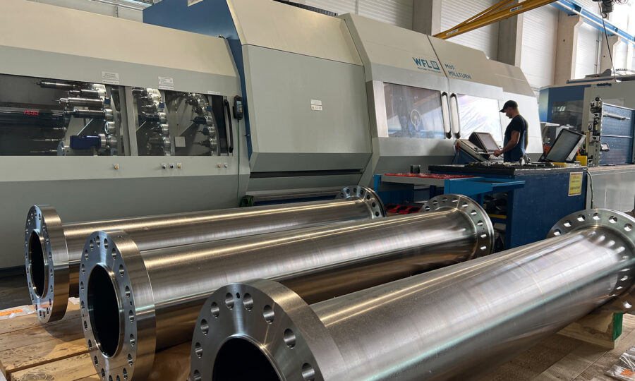 Große Metallzylinder mit Flanschen sind auf einer Palette vor einer industriellen CNC-Maschine in einer Fabrik aufgereiht. Ein Arbeiter bedient die Maschine, und der Arbeitsbereich wirkt sauber und ordentlich.