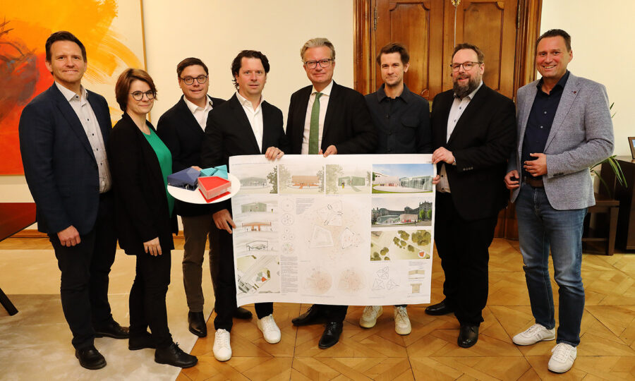 Acht Personen stehen in einem Innenraum in einem Halbkreis, drei von ihnen halten eine große Architekturtafel mit Zeichnungen, Diagrammen und Renderings. Alle tragen Geschäftskleidung oder elegante Freizeitkleidung.