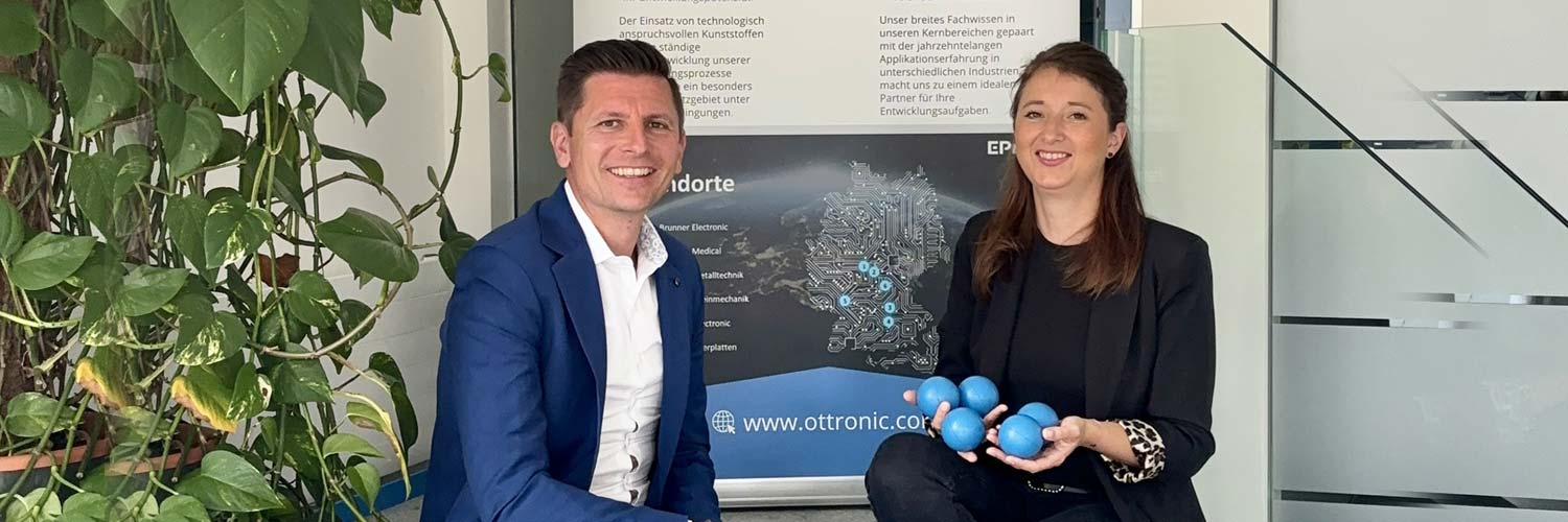Zwei Personen in Business-Kleidung lächeln in die Kamera. Die Frau hält mehrere blaue Bälle in ihren Händen. Sie sitzen drinnen in der Nähe einer Pflanze, vor einem großen Informationsplakat mit deutschem Text und einer Karte.