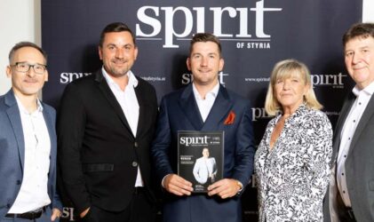 Fünf professionell gekleidete Personen stehen vor einem "spirit of styria"-Banner. Die Person in der Mitte hält ein Magazin mit demselben Branding. Sie lächeln und posieren für die Kamera.