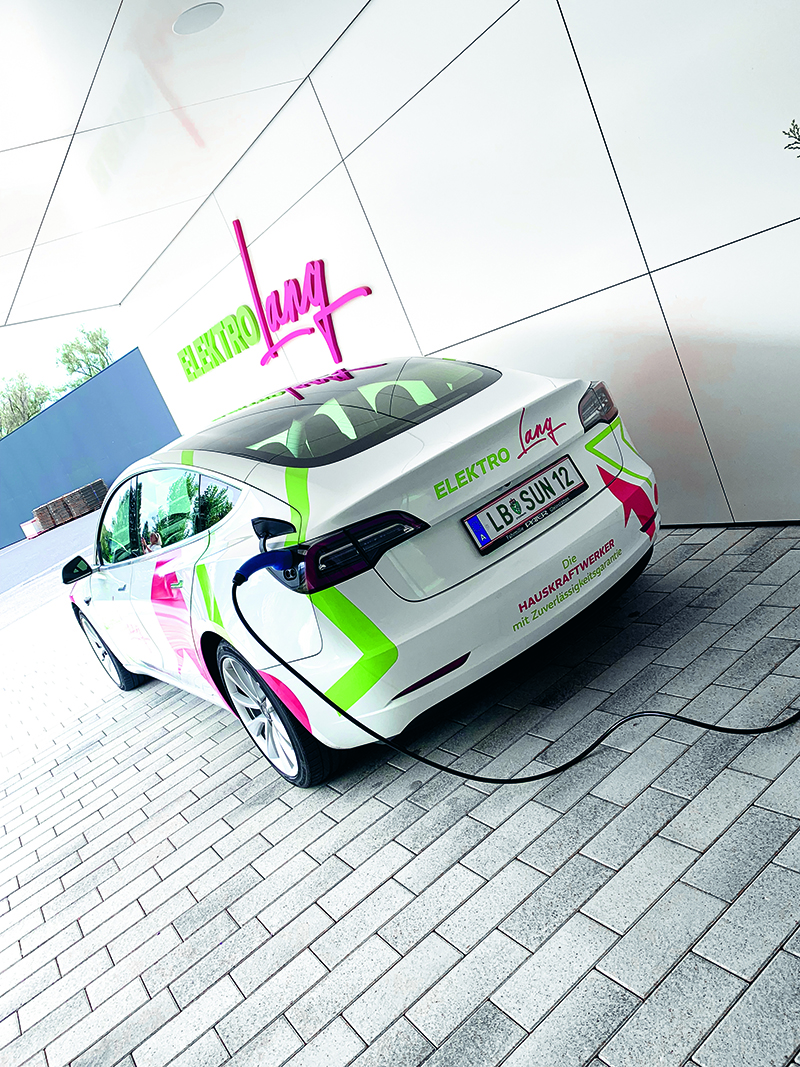 Ein weißes Elektroauto mit bunten grünen, rosafarbenen und gelben Grafiken parkt und lädt an einer modernen Station. Das Auto trägt das Wort "ELEKTRO" und ein deutsches Nummernschild, über dem eine Leuchtreklame "Lamy" zu sehen ist.