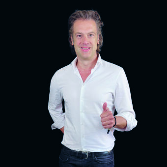 Ein Mann mit hellbraunem Haar, weißem Button-up-Hemd und dunkler Jeans steht vor einem schwarzen Hintergrund, lächelt und zeigt mit der rechten Hand den Daumen nach oben.