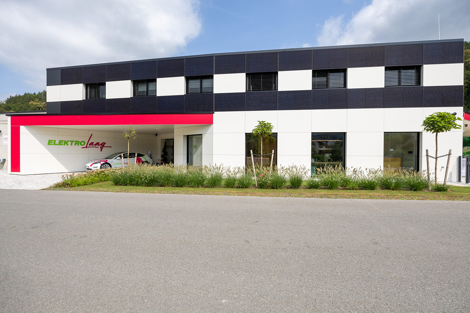 Ein modernes zweistöckiges Gebäude mit einer schwarz-weiß karierten Fassade, großen Fenstern und einem roten Akzent über dem Eingang. Auf dem Schild steht "ELEKTRO Lang". An der Fassade sind Gras und kleine Bäume gepflanzt.
