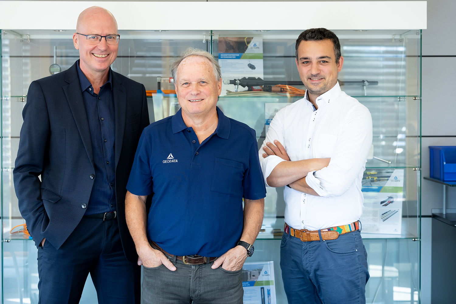 Drei Männer stehen lächelnd vor einer Vitrine mit Produkten und Informationsdisplays. Der Mann in der Mitte trägt ein blaues Polohemd, während die anderen in Geschäftskleidung sind. Der Hintergrund ist hell und modern.