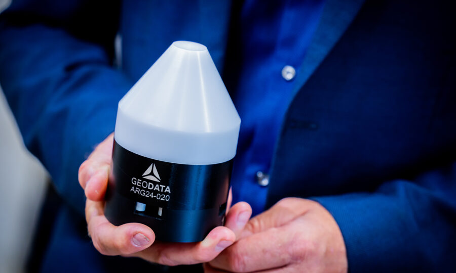 Eine Person in einem blauen Anzug hält ein kleines schwarz-weißes Gerät mit der Aufschrift "GEODATA ARG24-020" mit einem konischen Oberteil und einem zylindrischen Unterteil.