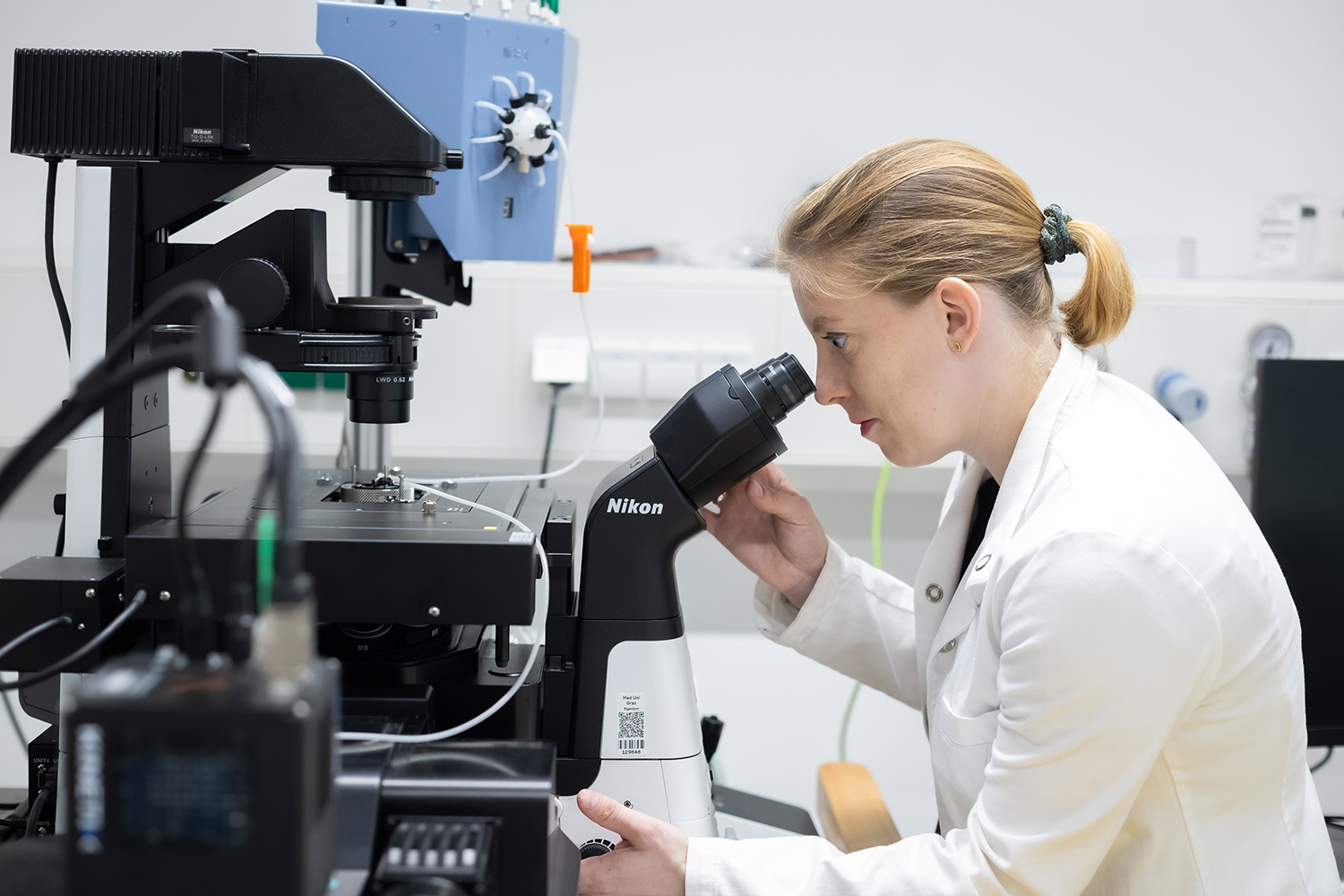 Eine Frau in einem weißen Laborkittel schaut durch ein Nikon-Mikroskop in einer Laborumgebung, umgeben von wissenschaftlichen Geräten und Instrumenten.