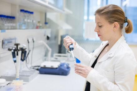 Ein Wissenschaftler in einem weißen Laborkittel benutzt eine Pipette, um in einem hellen Labor Flüssigkeit umzufüllen. Im Hintergrund sind wissenschaftliche Geräte und Flaschen auf den Regalen und der Theke zu sehen.