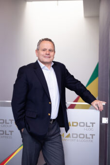 Ein Mann mittleren Alters in einem marineblauen Blazer und einem hellen Hemd steht selbstbewusst an eine Glaswand gelehnt, auf der der Firmenname "HADOLT" und bunte Diagonalstreifen zu sehen sind.
