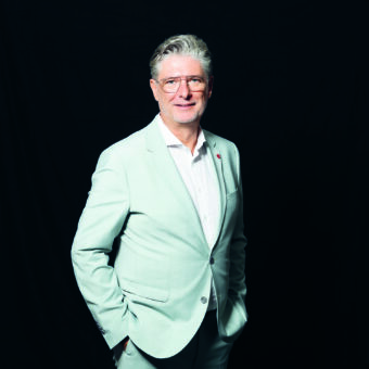Ein Mann mit grauem Haar und Brille, der einen hellen Anzug und ein weißes Hemd trägt, steht vor einem schlichten schwarzen Hintergrund und lächelt sanft, die Hände in den Taschen.