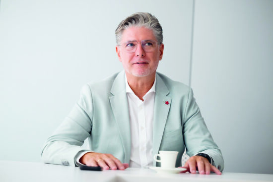 Ein Mann mit grauem Haar und Brille, hellem Anzug und weißem Hemd, sitzt an einem weißen Tisch mit Tasse und Untertasse und blickt leicht zur Seite in einem hellen, minimalistischen Raum.