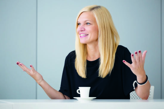 Eine lächelnde Frau mit langen blonden Haaren sitzt an einem Tisch mit einer kleinen weißen Kaffeetasse und gestikuliert mit beiden Händen. Sie trägt ein schwarzes Oberteil und eine Smartwatch und scheint in ein Gespräch vertieft zu sein.