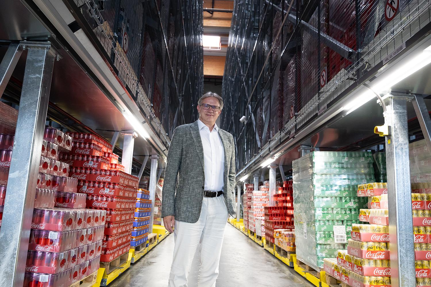 Ein Mann in einem hellen Anzug steht lächelnd in einem Lagergang, der mit Stapeln von Coca-Cola- und anderen Softdrink-Kisten gesäumt ist, wobei die Regale zu beiden Seiten hoch über ihn hinausragen.