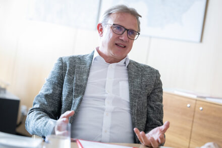 Ein Mann, der eine Brille und einen grauen Blazer trägt, sitzt an einem Schreibtisch und spricht in einem hellen Büro mit seinen Händen.