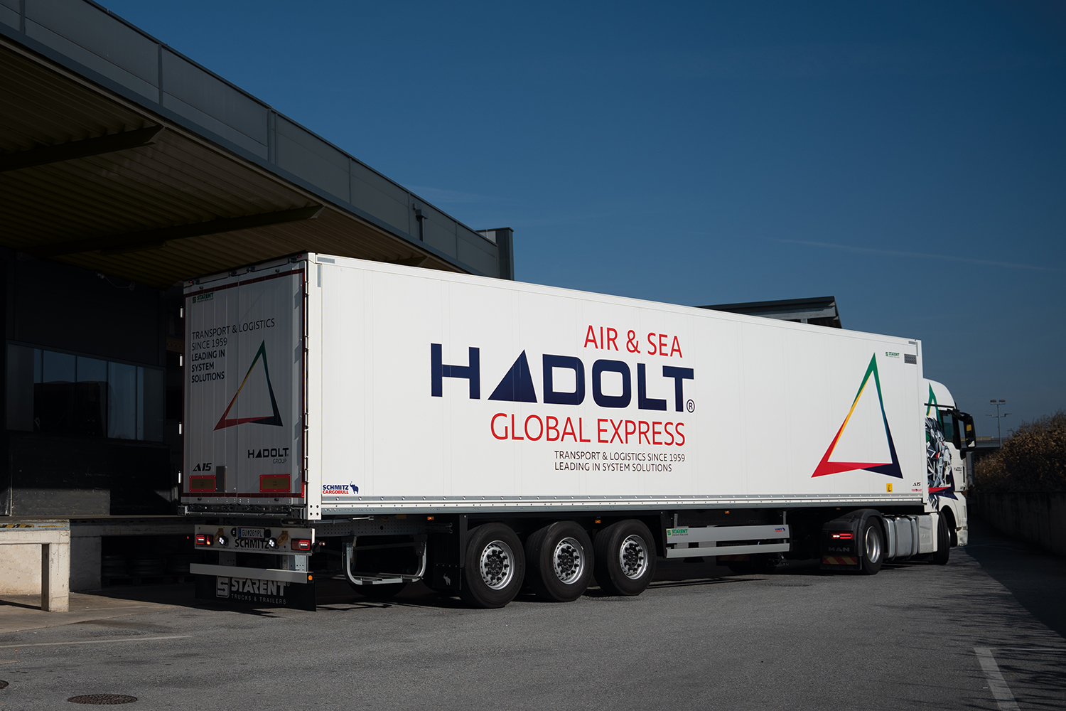 Ein weißer Hadolt Global Express-LKW ist an einer Laderampe geparkt. Der Lkw trägt das Firmenlogo und den Slogan "Air & Sea Global Express" auf der Seite, mit einer bunten dreieckigen Grafik neben dem Text.