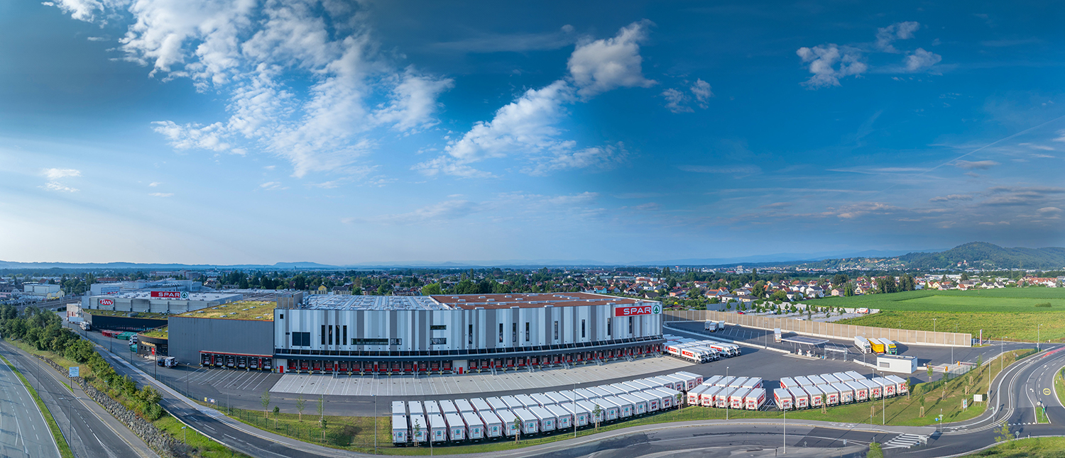 Ein großes SPAR-Lager und -Vertriebszentrum mit Reihen von Laderampen und geparkten Lastwagen, eingebettet in eine halbstädtische Landschaft unter einem blauen Himmel mit vereinzelten Wolken.
