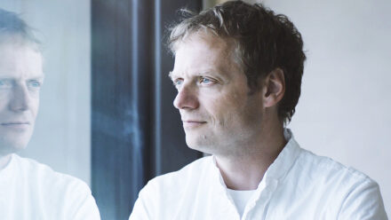 Ein Mann mit hellbraunem Haar, der ein weißes Hemd trägt, steht in einem Haus und schaut nachdenklich aus dem Fenster. Sein Spiegelbild ist im Glas neben ihm zu sehen.