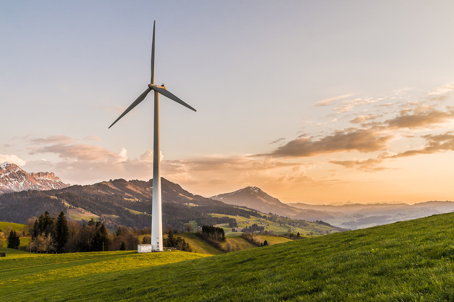Eine große Windturbine steht auf einem grasbewachsenen Hügel bei Sonnenuntergang, mit Bergen und verstreuten Bäumen im Hintergrund unter einem teilweise bewölkten Himmel.