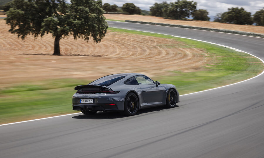 Ein schnittiger grauer Sportwagen rast um eine Kurve auf einer Rennstrecke, mit unscharfen Bewegungen im Vordergrund und trockenen Feldern und Bäumen im Hintergrund unter einem teilweise bewölkten Himmel.