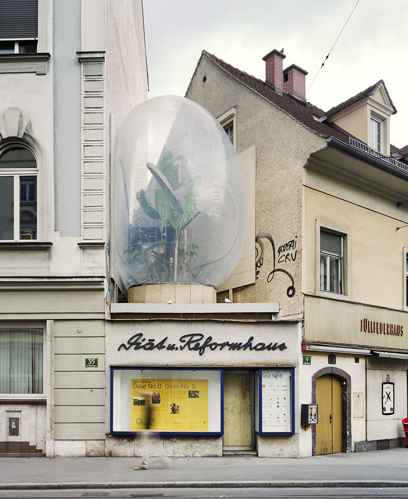 Ein Schaufenster mit der Aufschrift "Gädu Reformhaus" hat eine große blasenartige Kuppel auf dem Dach, die hohe Grünpflanzen umschließt. Das Gebäude befindet sich zwischen zwei anderen Gebäuden an einer Stadtstraße.
