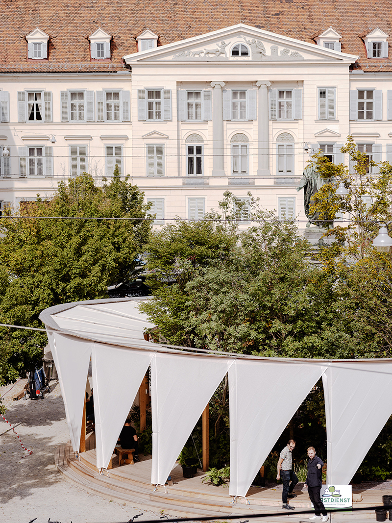 Ein runder Holzpavillon mit weißen Stoffbahnen steht inmitten von Bäumen vor einem großen, historischen Gebäude mit hohen Fenstern und einem roten Ziegeldach. Zwei Personen stehen am Eingang des Pavillons, und auf einem Schild steht "StadtErnst".