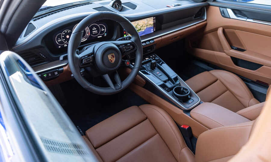 Das Bild zeigt den Innenraum eines modernen Porsche-Sportwagens mit einem schwarzen Lenkrad mit Porsche-Logo, digitalen Anzeigen auf dem Armaturenbrett, hellbraunen Ledersitzen und einer eleganten, hochtechnisierten Mittelkonsole.