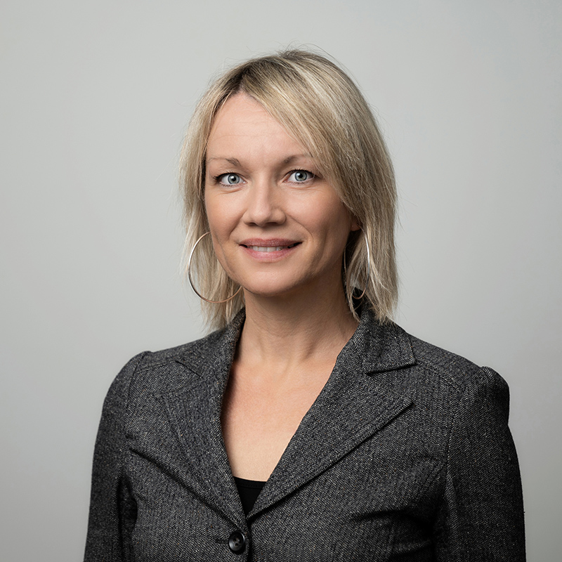 Eine Frau mit blondem Haar, die einen grauen Blazer und große Ohrringe trägt, lächelt in die Kamera vor einem schlichten hellgrauen Hintergrund.