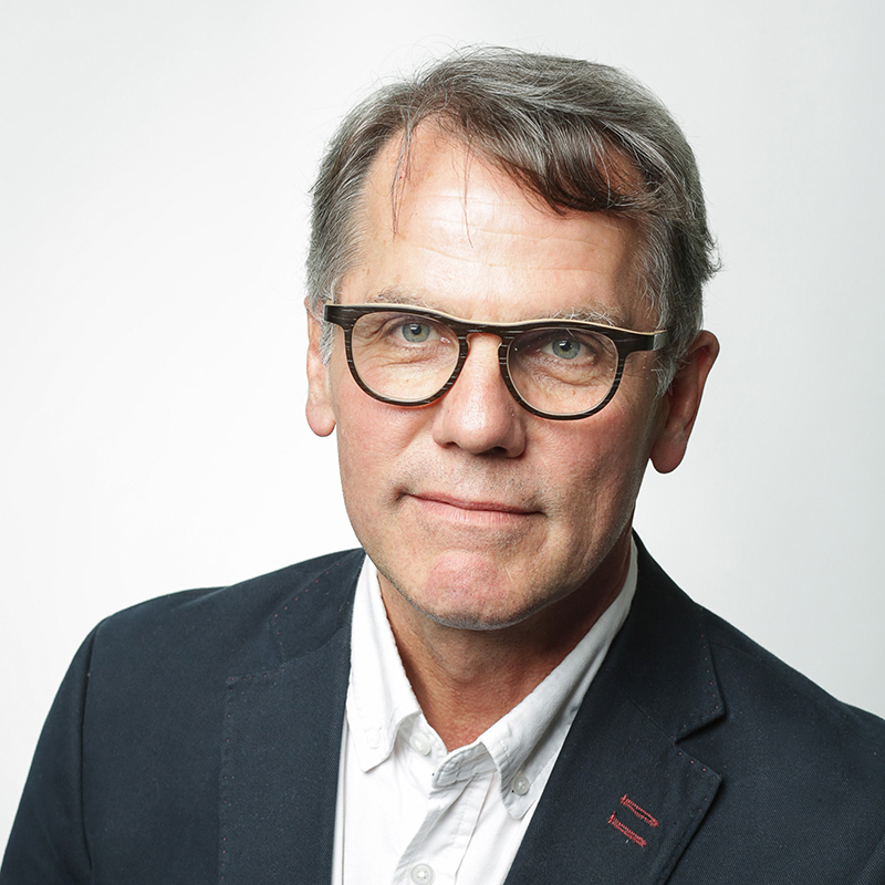 Ein Mann mittleren Alters mit kurzen grauen Haaren, der eine Brille, ein weißes Hemd und einen dunklen Blazer trägt, posiert vor einem schlichten hellen Hintergrund.