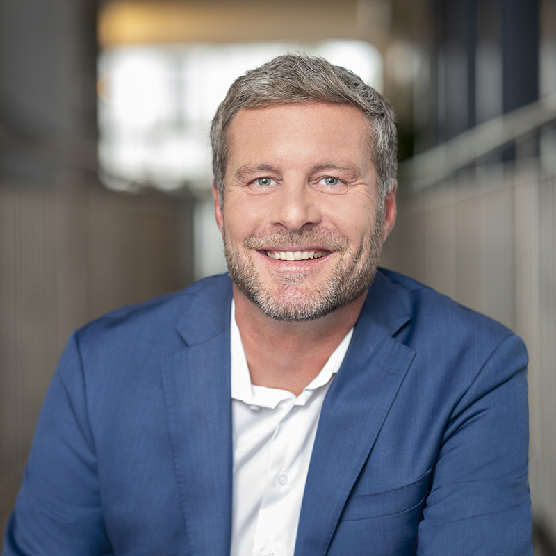 Ein lächelnder Mann mit kurzen grauen Haaren und einem Bart, der einen blauen Blazer über einem weißen Hemd trägt, sitzt in einem hell erleuchteten Innenraum mit unscharfem Hintergrund.