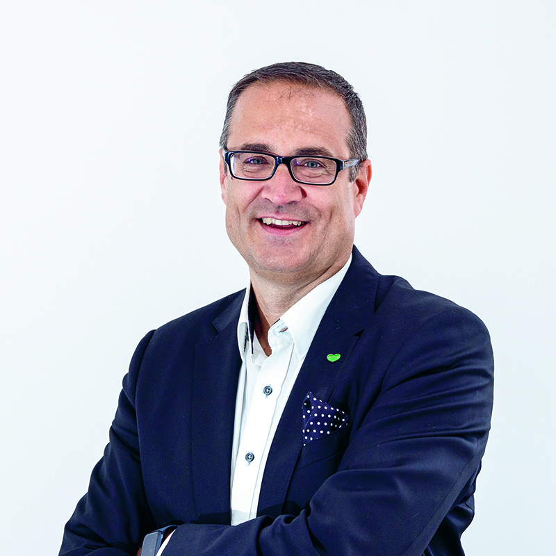 Ein Mann mit Brille, weißem Hemd und dunklem Blazer mit gepunktetem Einstecktuch steht lächelnd mit verschränkten Armen vor einem schlichten weißen Hintergrund.