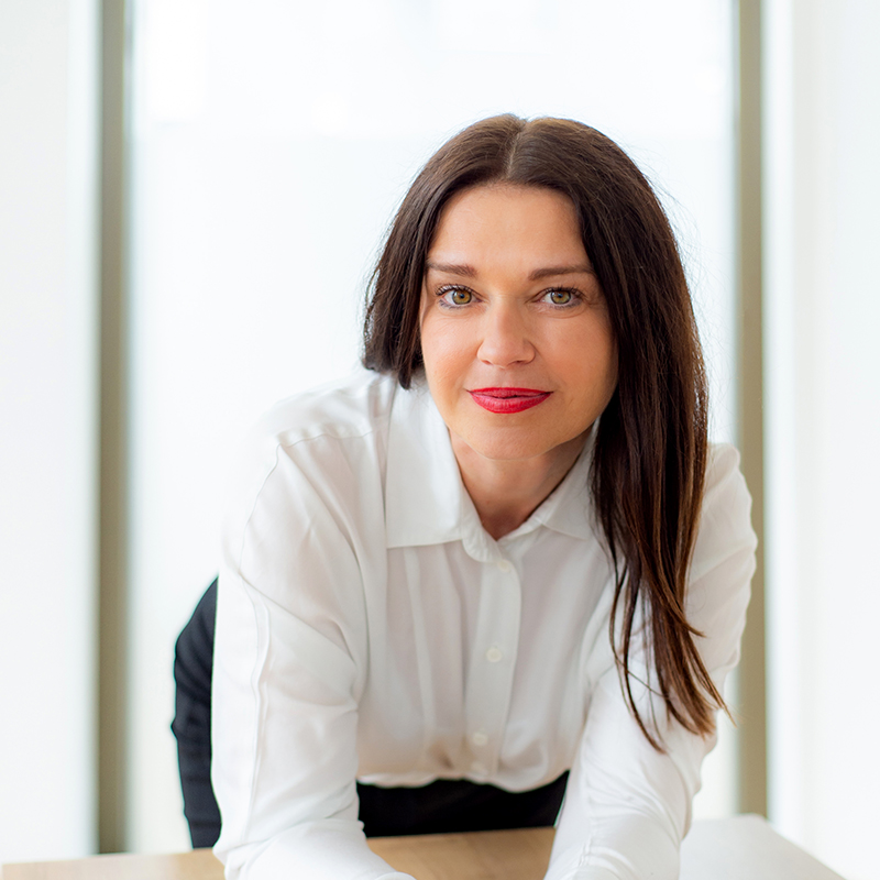 Eine Frau mit langen dunklen Haaren und weißem Button-up-Hemd lehnt sich leicht lächelnd an einen Holztisch, der Hintergrund ist hell und sanft beleuchtet.