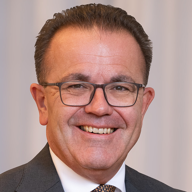 Ein Mann mittleren Alters mit kurzen braunen Haaren, Brille und Anzug lächelt in die Kamera vor einem unscharfen hellen Hintergrund.