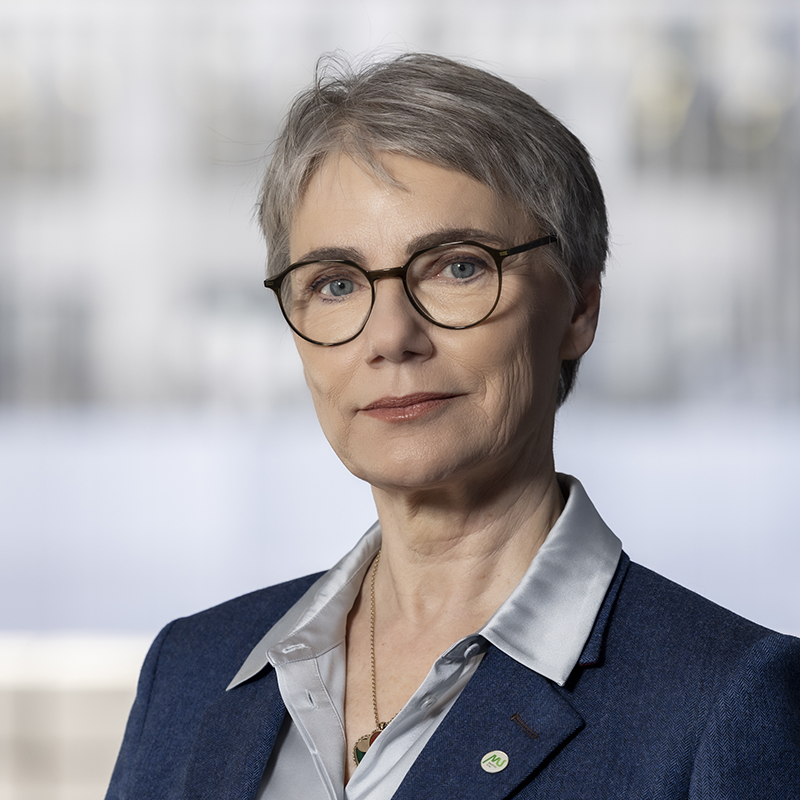 Eine Frau mit kurzen grauen Haaren und Brille trägt einen blauen Blazer über einem hellen Hemd und steht vor einem unscharfen Hintergrund. Sie blickt selbstbewusst in die Kamera.
