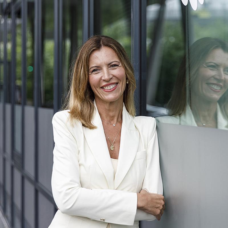 Eine Frau mit schulterlangen hellbraunen Haaren in einem weißen Blazer steht lächelnd mit verschränkten Armen an eine Glaswand gelehnt, in der sich ihr Spiegelbild spiegelt. Im Hintergrund ist draußen eine Grünanlage zu sehen.