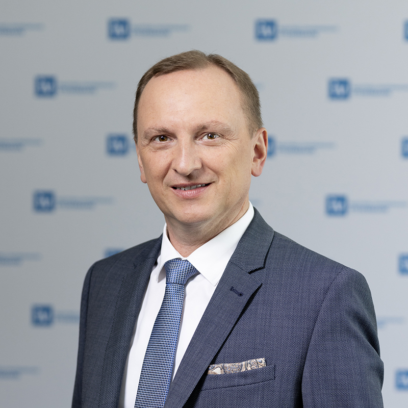 Ein Mann in blauem Anzug und Krawatte lächelt in die Kamera und steht vor einem verschwommenen Hintergrund mit sich wiederholenden blauen und weißen Logos.
