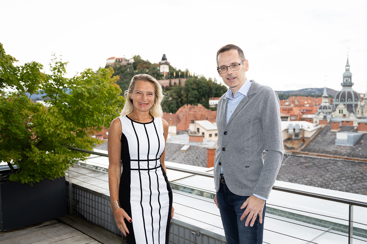 Eine Frau in einem schwarz-weißen Kleid und ein Mann in einem grauen Pullover stehen auf einer Terrasse mit einer Stadtlandschaft, roten Dächern und einem Uhrenturm im Hintergrund. Auf der linken Seite steht ein belaubter Baum.