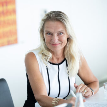 Eine Frau mit langen blonden Haaren in einem ärmellosen weiß-schwarzen Kleid sitzt lächelnd an einem Tisch und hält einen Stift mit Papieren und ein Glas Wasser vor sich. Der Hintergrund ist hell und leicht unscharf.