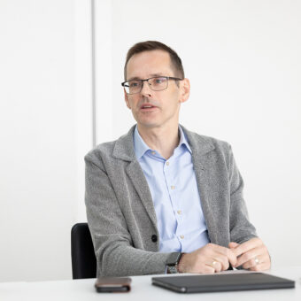 Ein Mann, der eine Brille, einen grauen Blazer und ein hellblaues Hemd trägt, sitzt an einem Tisch mit einem Notebook und einem Telefon vor sich und spricht und gestikuliert mit seinen Händen in einer hellen, modernen Umgebung.