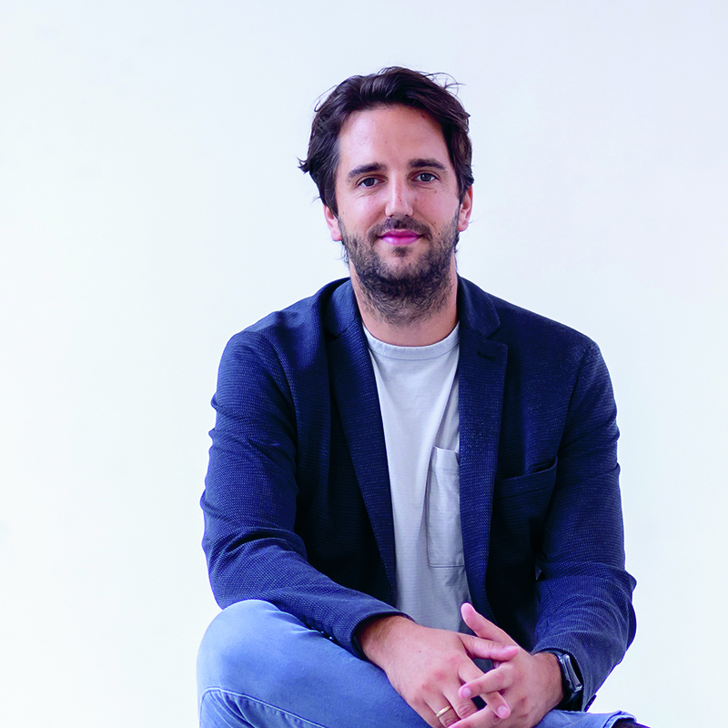 Ein Mann mit dunklem Haar und Bart, der einen marineblauen Blazer über einem weißen T-Shirt und blaue Jeans trägt, sitzt lässig vor einem schlichten weißen Hintergrund und schaut mit einem leichten Lächeln in die Kamera.