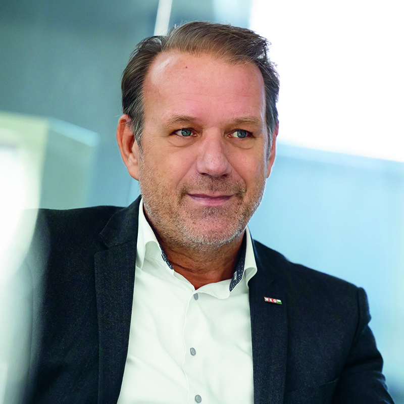 Ein Mann mittleren Alters mit kurzen braunen Haaren und gestutztem Bart, der ein weißes Hemd und eine dunkle Anzugsjacke trägt, sitzt in einem Raum und schaut leicht zur Seite, wobei er leicht lächelt.
