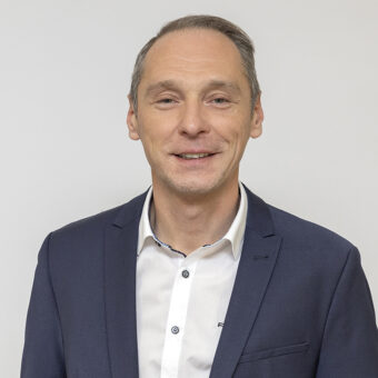 Ein Mann mit kurzen, hellbraunen Haaren, der eine dunkelblaue Anzugsjacke und ein weißes Hemd mit Knöpfen trägt, lächelt und steht vor einem schlichten, hellen Hintergrund.