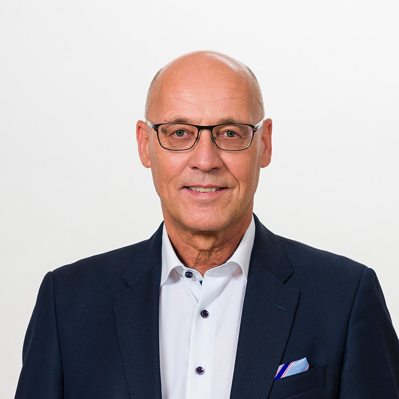 Ein Mann mit Glatze, Brille, dunkler Anzugjacke und weißem Hemd mit Einstecktuch, der vor einem schlichten weißen Hintergrund leicht lächelt.