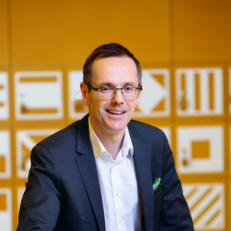 Ein Mann, der eine Brille, einen dunklen Anzug und ein weißes Hemd trägt, lächelt in die Kamera. Er steht vor einer gelb-orangenen Wand mit geometrischen weißen Mustern.