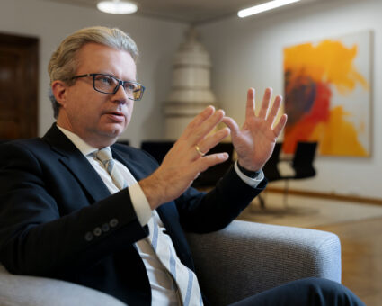 Ein Mann in Anzug und Krawatte sitzt auf einem grauen Stuhl und gestikuliert mit beiden Händen, während er spricht. Im Hintergrund sind abstrakte Kunstwerke an der Wand und ein Modell eines weißen Gebäudes zu sehen.