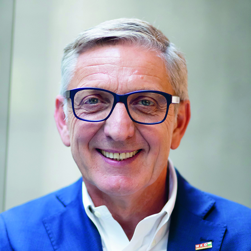 Ein lächelnder älterer Mann mit kurzen grauen Haaren und blauer Brille, der eine blaue Anzugsjacke und ein weißes Hemd mit Kragen trägt, steht vor einem sanft verschwommenen Hintergrund.