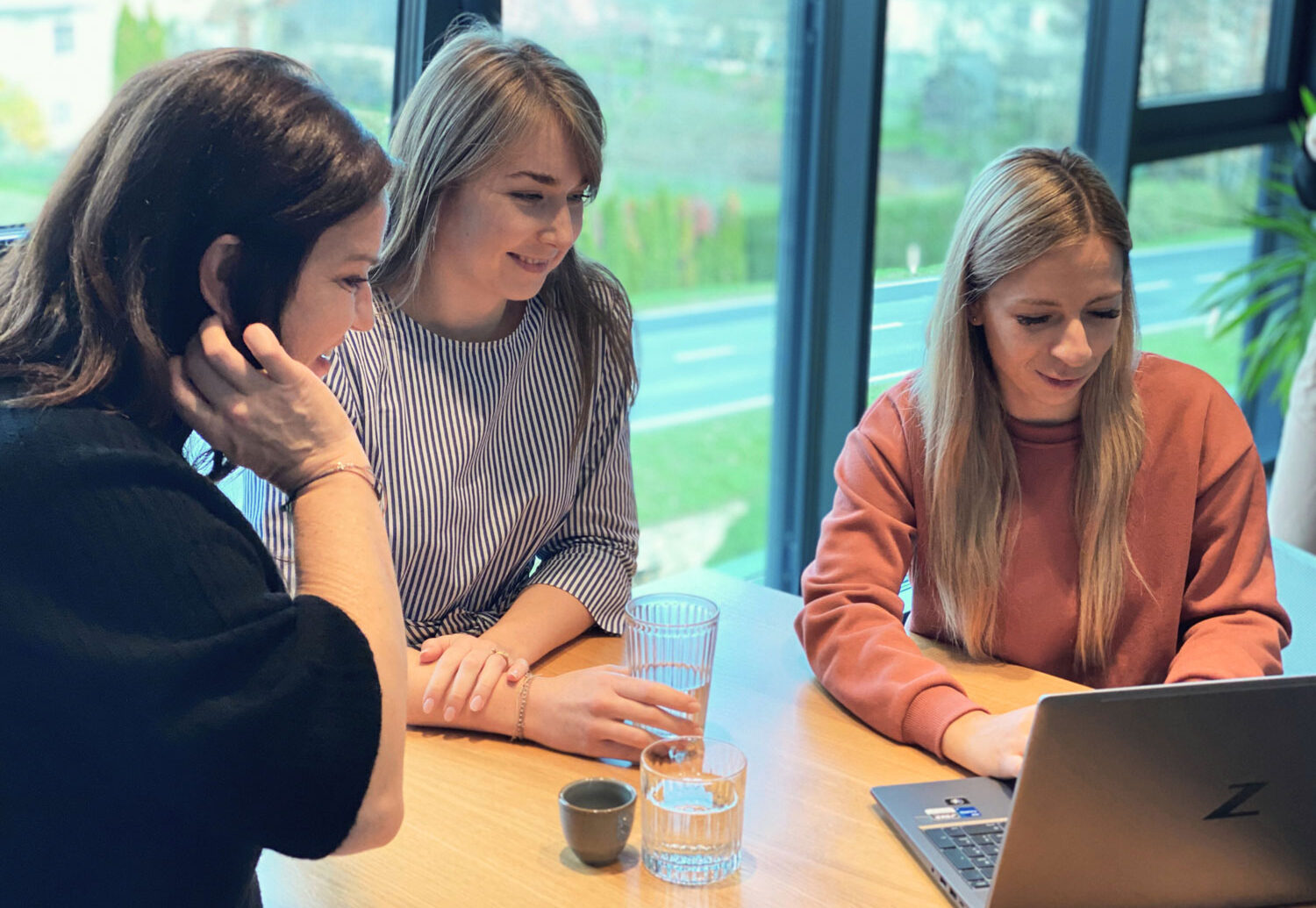 Drei Frauen sitzen an einem Tisch mit Getränken, lächeln und schauen gemeinsam auf einen Laptop-Bildschirm. Große Fenster hinter ihnen zeigen einen verschwommenen Blick auf Bäume und eine Straße draußen.