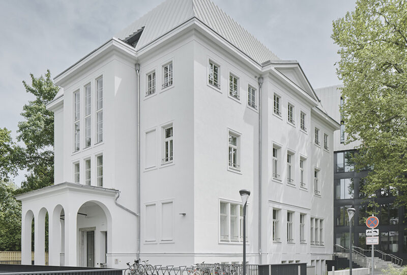 Ein großes weißes Gebäude mit hohen Fenstern, einem modernen Metalldach und einem bogenförmigen Eingang. Vor dem Gebäude sind Fahrräder abgestellt, und im Hintergrund sind Bäume und eine moderne Glaskonstruktion zu sehen.