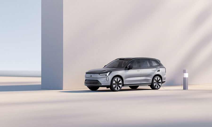 Ein silberner Elektro-Geländewagen parkt neben einer modernen weißen Wand, angeschlossen an eine Ladestation in einer minimalistischen, sonnenbeschienenen Umgebung mit weichen Schatten.