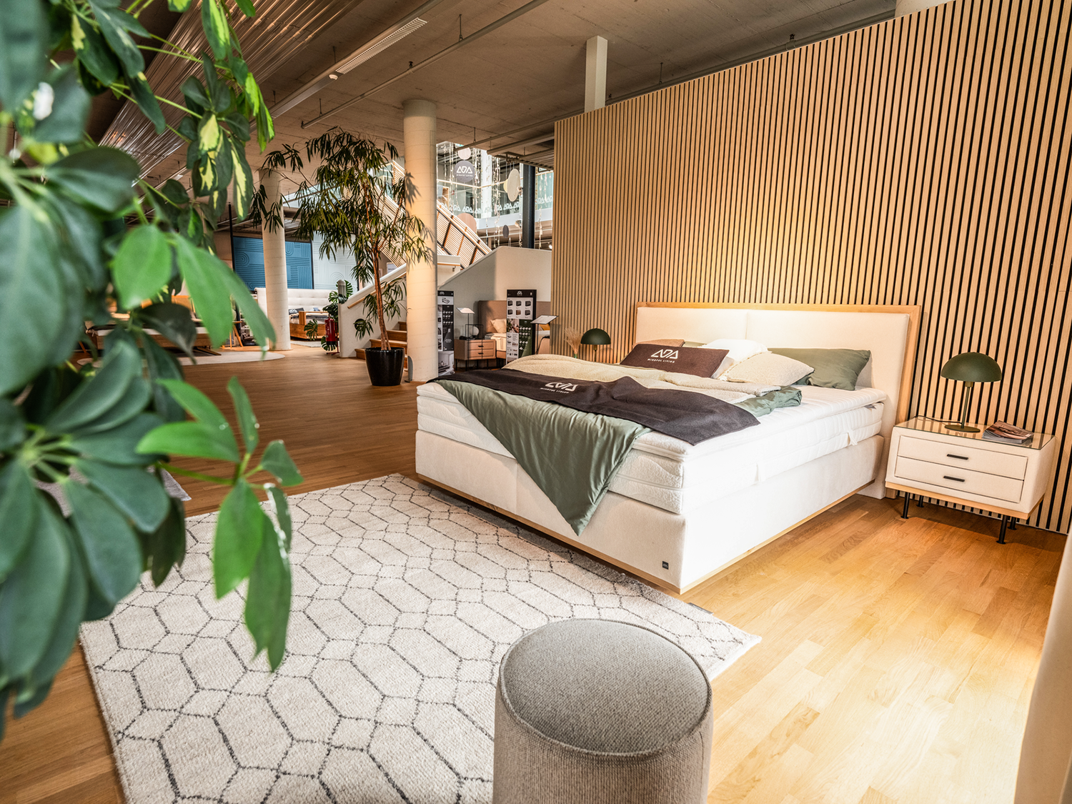 Ein modernes Schlafzimmer mit einem großen Bett, grüner und weißer Bettwäsche, zwei Nachttischen mit Lampen, einem geometrischen Teppich, Topfpflanzen und Holzfußböden in einem geräumigen, offenen Raum mit Holzlattenwänden.