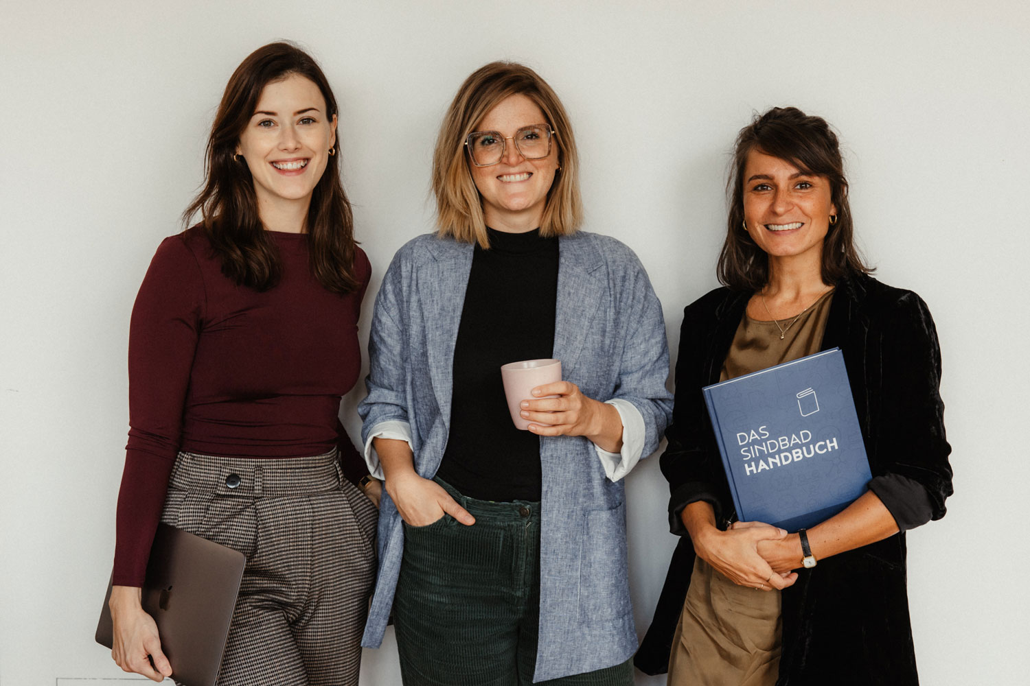 Drei Frauen stehen lächelnd zusammen. Eine hält einen Laptop, eine andere eine rosa Tasse und die dritte ein blaues Buch mit der Aufschrift "Das Sindda Handbuch". Sie sind professionell gekleidet und stehen vor einem schlichten weißen Hintergrund.